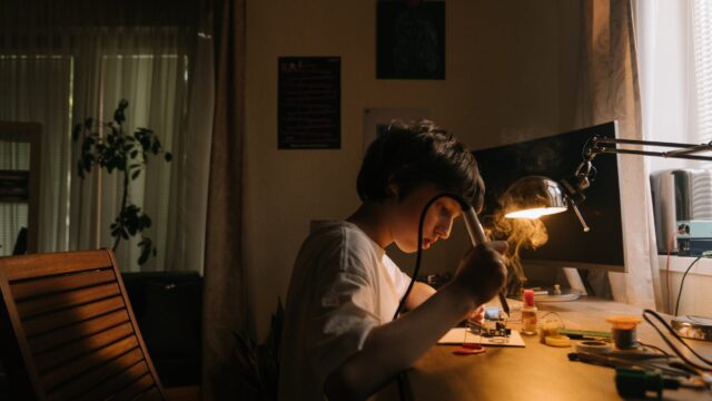A child holds a soldering iron while looking at a soldering project. The smoke from soldering is visible in the bright light from a lamp on the work surface. Various electronics tools are scattered around the work surface.